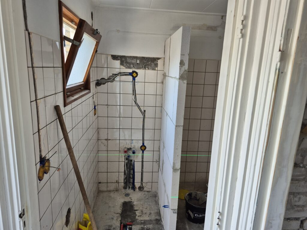 Oude doucheruimte met wit tegelwerk en leidingwerk in het zicht, klaar voor verbouwing